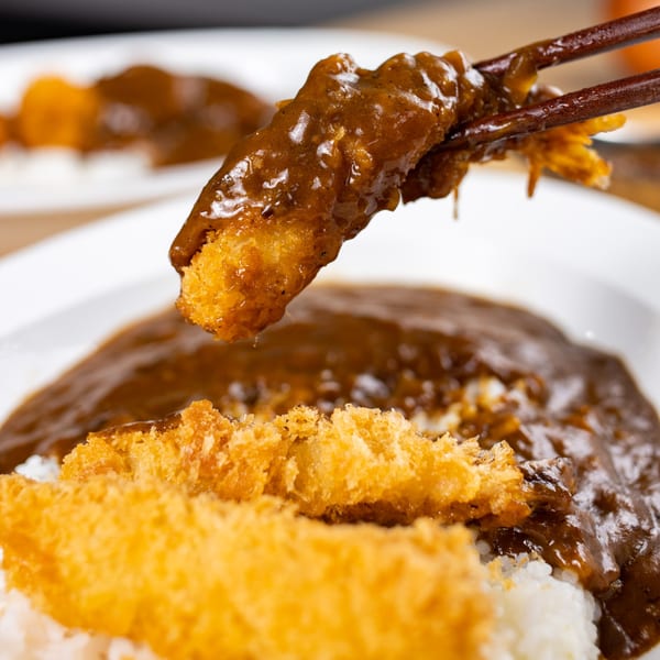 Chopsticks holding a piece of fried shrimp coated in curry sauce, above a plate of rice and curry.