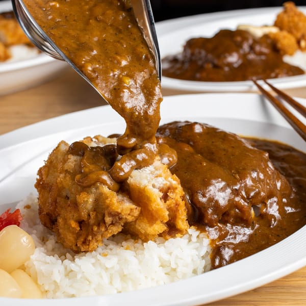 Curry sauce being poured over crispy fried chicken katsu and white rice with pickled vegetables.